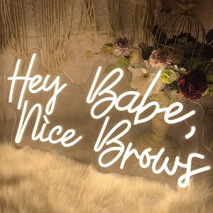 May include: White neon sign with the text "Hey Babe, Nice Brows" on a clear acrylic background. The sign is sitting on a white fluffy surface.