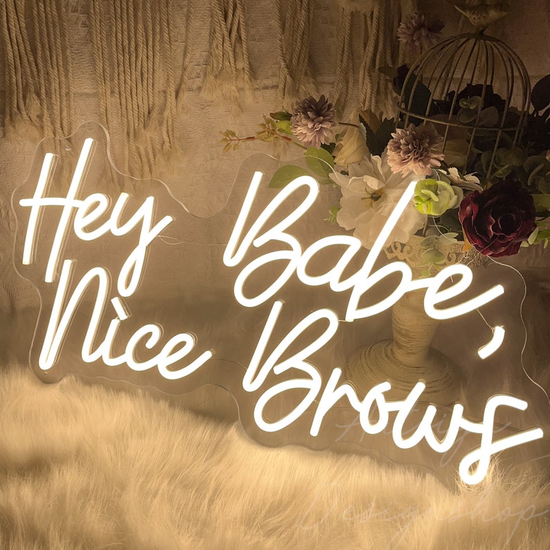 May include: White neon sign with the text "Hey Babe, Nice Brows" on a clear acrylic background. The sign is sitting on a white fluffy surface.