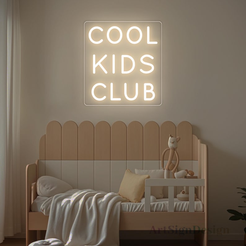 Op de afbeelding: Een wit neonbord met de woorden "COOL KIDS CLUB" op een witte muur boven een houten bed met een wit en beige hoofdbord.