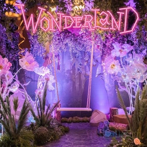 May include: A whimsical wonderland scene with a neon sign that reads "WONDERLAND" and a white swing hanging from a purple floral backdrop. The scene is decorated with large, white paper flowers and a purple, leafy forest.