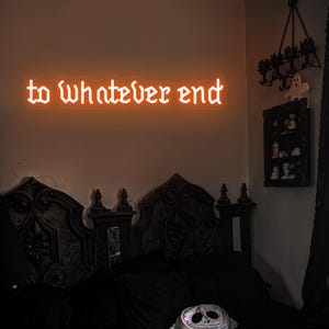 May include: A neon sign with the text "to whatever end" in a gothic font, mounted on a wall above a dark wooden headboard with a decorative design. The sign is lit up in a warm orange color.