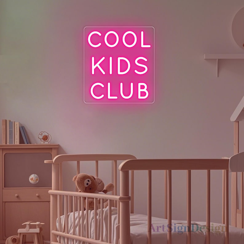 Op de afbeelding: Een roze neonbord met de tekst "COOL KIDS CLUB" op een witte achtergrond. Het bord hangt aan een muur in een kinderkamer met een wieg en een teddybeer.