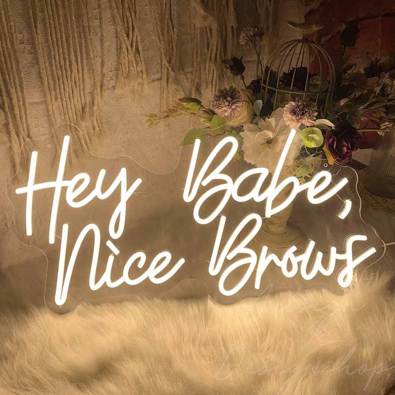 May include: White neon sign with the text "Hey Babe, Nice Brows" on a white fluffy surface.
