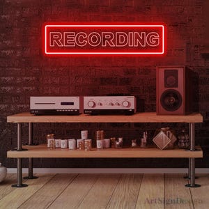 Op de afbeelding: Een rechthoekig neonbord met een felrode rand en het woord RECORDING in witte hoofdletters. Het bord is gemonteerd op een donkere bakstenen muur boven een houten rek met twee niveaus, gevuld met audioapparatuur, kleine witte bekers en glaswerk.