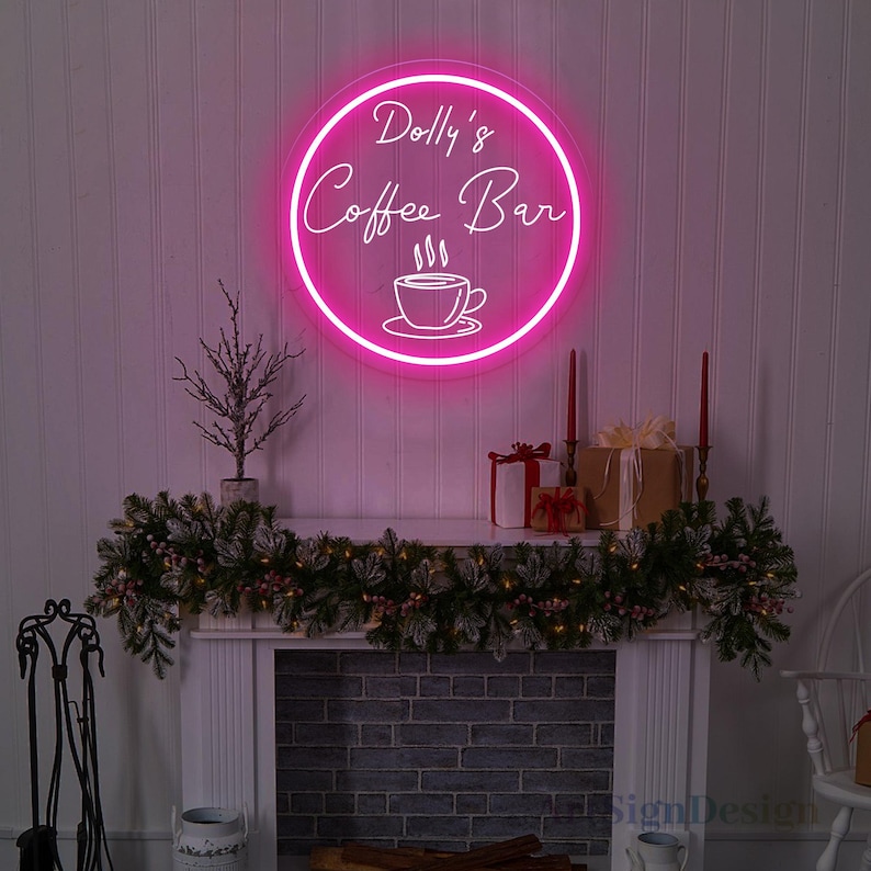 May include: A pink neon sign with a white outline that reads "Dolly's Coffee Bar" with a stylized coffee cup image below the text. The sign is hanging on a white wall above a fireplace mantel decorated with a green garland.