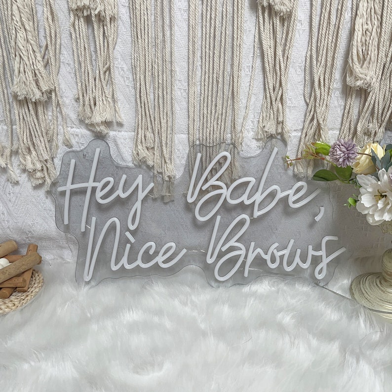 May include: White neon sign with the text "Hey Babe, Nice Brows" on a clear acrylic background.