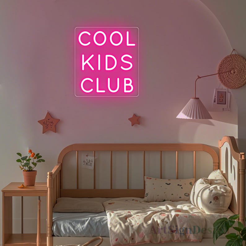 Op de afbeelding: Een roze neon-bord met de tekst "COOL KIDS CLUB" hangt aan een muur boven een wit en bruin dagbed met witte beddengoed en kussens.