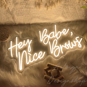 May include: White neon sign with the text "Hey Babe, Nice Brows" on a white fluffy surface.