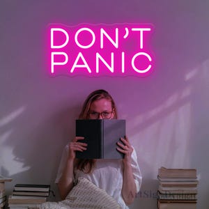 Könnte beinhalten: Ein pinkfarbenes Neon-Schild mit dem Text "DON'T PANIC" an einer weißen Wand. Eine Person sitzt vor dem Schild und hält ein Buch vor ihrem Gesicht.