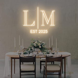 Può includere: Un'insegna al neon con le lettere "L" e "M" e la scritta "EST 2025". È posta sopra un tavolo allestito per un ricevimento di nozze, con candele, fiori e posate. Le sedie circondano il tavolo.