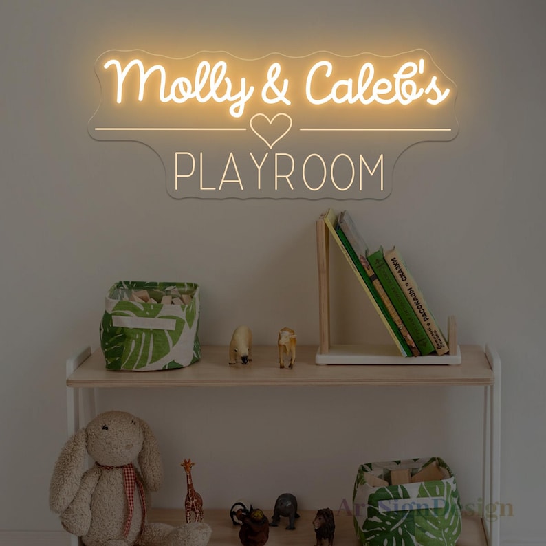 May include: A neon sign with the text "Molly & Caleb's PLAYROOM" in a playful font. Below, a wooden shelf displays a stuffed bunny, toy animals, books, and storage bins with a green leaf pattern.