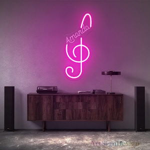 Op de afbeelding: Een roze neon-bord met een vioolsleutel en de naam "Amanda" in cursief schrift. Het bord is gemonteerd op een muur boven een houten kast met twee luidsprekers aan beide kanten.