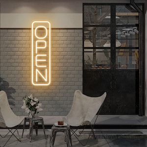 May include: A neon sign with the word "OPEN" in a vertical rectangular frame is mounted on a brick wall. Two white butterfly chairs and small metal tables are in front of the wall. A vase of white flowers and a small cake are on the tables.