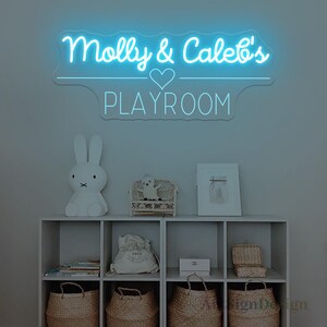 May include: A neon sign in light blue illuminates the words "Molly & Caleb's PLAYROOM" above a shelf. The shelf displays a white bunny lamp, a small wicker chair with a doll, a framed picture, and woven baskets. The sign is a decorative element for a playroom.