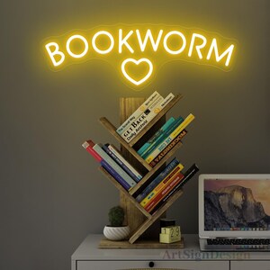 May include: A wooden bookshelf shaped like a tree with books on each shelf. The bookshelf is on a white table with a laptop and a small potted plant. A yellow neon sign with the words "BOOKWORM" and a heart is above the bookshelf.