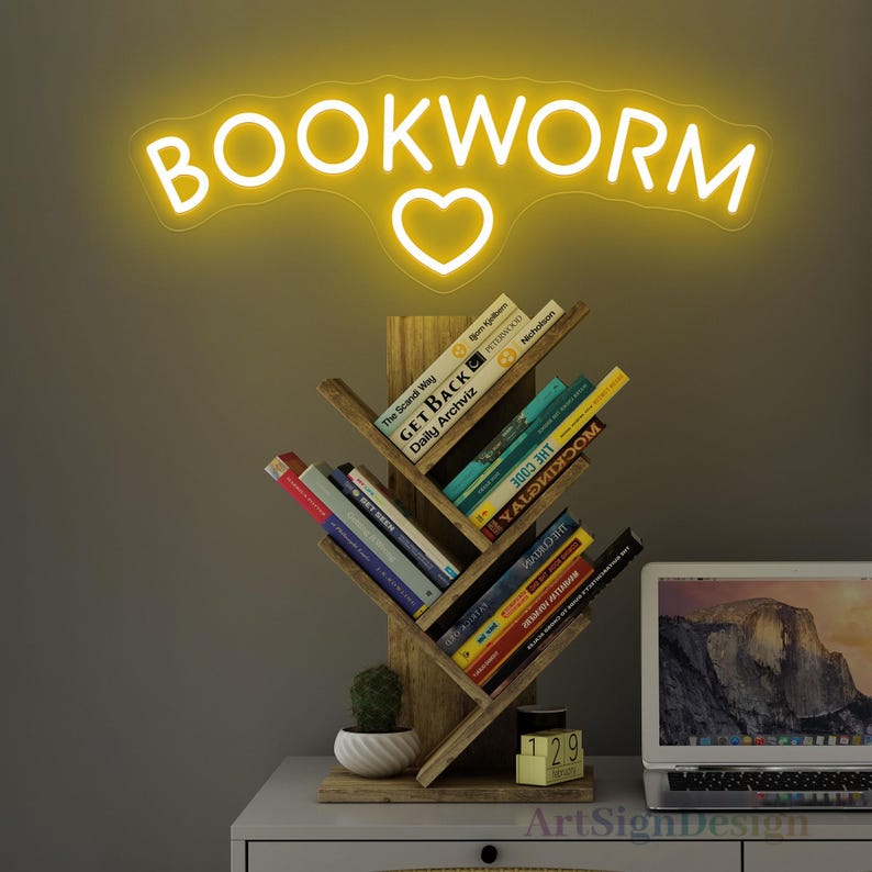 May include: A wooden bookshelf shaped like a tree with books on each shelf. The bookshelf is on a white table with a laptop and a small potted plant. A yellow neon sign with the words "BOOKWORM" and a heart is above the bookshelf.