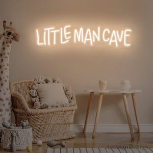 Op de afbeelding: Een neonbord voor aan de muur met de tekst "LITTLE MAN CAVE" in een warm, wit schijnend licht. Het bord heeft een op maat gemaakte, golvende acryl achterplaat die de contouren van de letters volgt en zorgt voor een zachte verlichting.