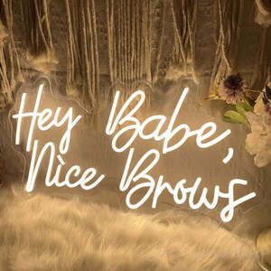 May include: White neon sign with the text "Hey Babe, Nice Brows" in a cursive font.