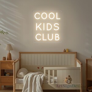 Op de afbeelding: Een wit neon-bord met de woorden "COOL KIDS CLUB" op een witte achtergrond. Het bord hangt aan een muur boven een houten wieg.