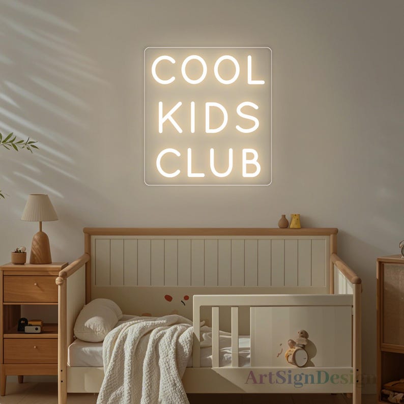 Op de afbeelding: Een wit neon-bord met de woorden "COOL KIDS CLUB" op een witte achtergrond. Het bord hangt aan een muur boven een houten wieg.
