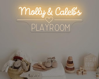 Puede incluir: Un letrero de neón con el texto "Molly & Caleb's PLAYROOM" en una fuente lúdica. Un símbolo de corazón está centrado entre los nombres y la palabra "PLAYROOM". Debajo, una exhibición de artículos para bebés, incluidos juguetes y zapatos.