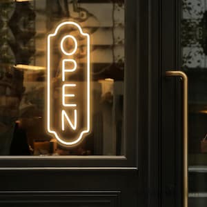 May include: A white neon sign with the word "OPEN" in a decorative frame, displayed in a storefront window.