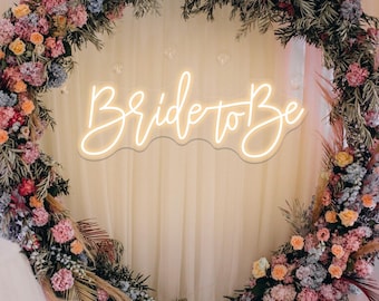 Pode incluir: Uma placa de néon branca com o texto "Bride to Be" em uma fonte cursiva. A placa está pendurada na frente de um arco floral feito de flores rosa, laranja e azuis.