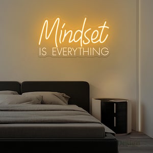 Può includere: Un'insegna al neon con le parole "Mindset IS EVERYTHING" in un carattere corsivo, che brilla con una calda luce gialla. L'insegna è montata su un muro sopra un letto con lenzuola grigie e una testiera nera. Un comodino è sulla destra.