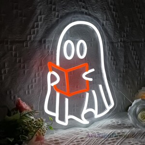 Op de afbeelding: Een wit neon-bord met een geest die een boek leest. De geest heeft twee grote ogen en een boek met een oranje gloed. Het bord is op een donkere achtergrond.