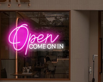 May include: Pink neon sign with the word "Open" and the phrase "Come on in" in white lettering. The sign is hanging in a window of a cafe.