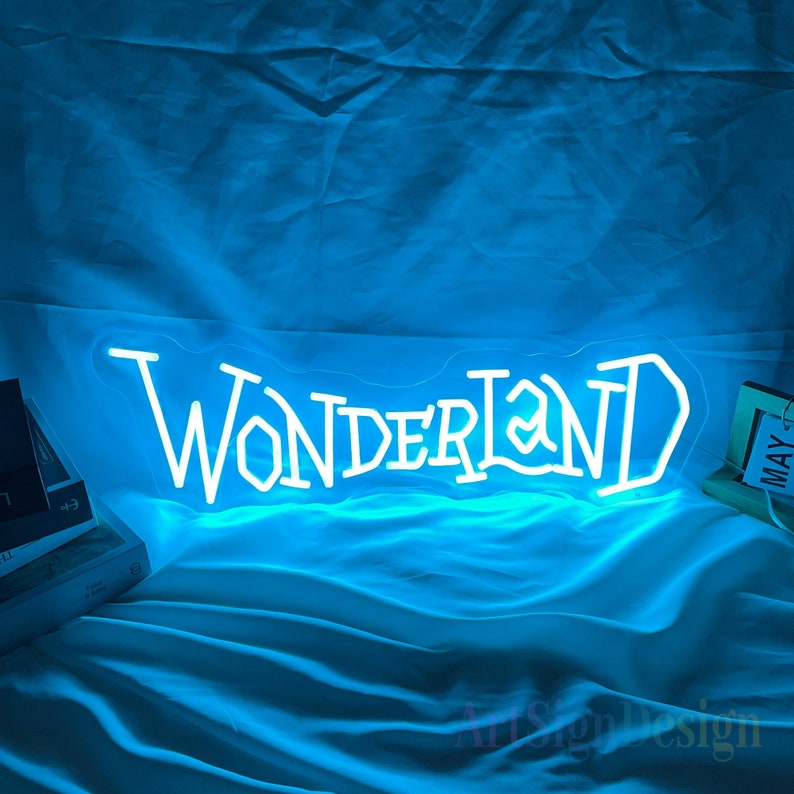 May include: White neon sign with the word "WONDERLAND" in a stylized font. The sign is lit up with a bright blue light and is sitting on a blue fabric surface.