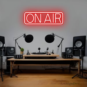 Op de afbeelding: Een rechthoekig neonbord met een rode rand en de tekst ON AIR in witte hoofdletters. Het bord is aan een muur bevestigd boven een houten bureau met audio-opnameapparatuur, waaronder twee microfoons, luidsprekers en een draaitafel.