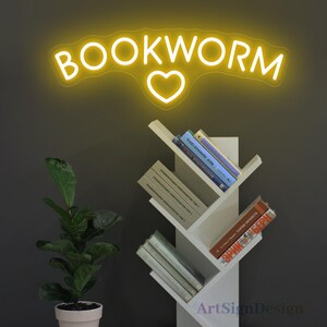 May include: Yellow neon sign with the word "BOOKWORM" and a heart in the middle. The sign is mounted on a wall above a white bookshelf with books.