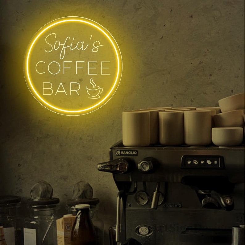 May include: A yellow neon sign with the text "Sofia's Coffee Bar" and a coffee cup icon. The sign is mounted on a wall behind a coffee machine.