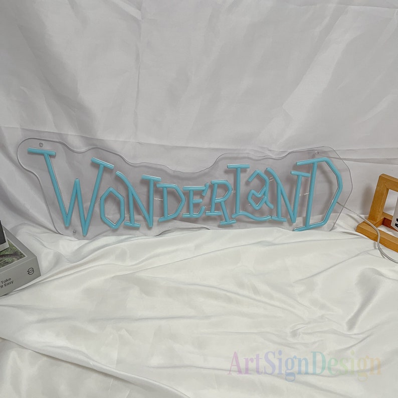 May include: A light blue neon sign with the word "WONDERLAND" in a whimsical font. The sign is mounted on a clear acrylic backing.