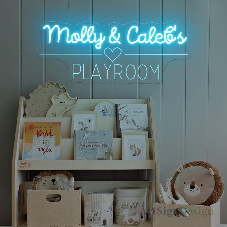 May include: A neon sign in blue script reads "Molly & Caleb's PLAYROOM" above a wooden bookshelf filled with books and decorative items. The shelf holds a hedgehog and lion plush toys, and decorative storage containers. The overall theme is children's decor.