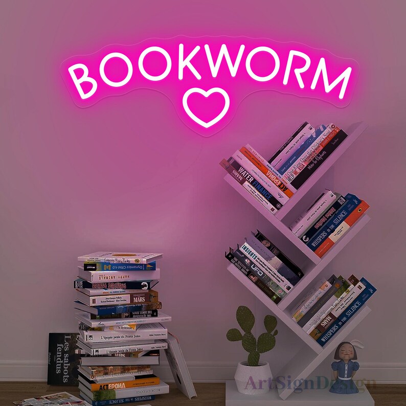 May include: Pink neon sign with the word "BOOKWORM" and a heart in the middle. The sign is mounted on a white wall above a white bookshelf with books.