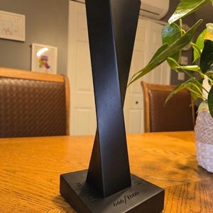 Peut inclure: Une sculpture noire et géométrique avec un design torsadé, posée sur une base rectangulaire. La base porte le mot "PRESENCE" et le numéro "666/1000". La sculpture est exposée sur une table en bois.