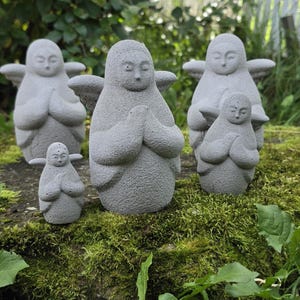 May include: A collection of four gray stone-like angel figurines with textured surfaces. Each figure features a rounded body, small wings, and hands pressed together in a prayer position. The set includes two larger figures and two smaller figures.