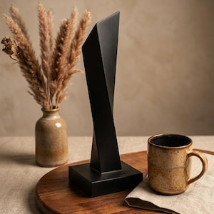 May include: A matte black sculptural object with a twisted, geometric vertical form standing on a square base. The piece features sharp, angular edges and a smooth, solid finish, positioned upright as a decorative item on a flat surface.