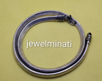 Può includere: Una collana color argento con un design a forma di serpente, dotata di chiusura a fermaglio. La collana circolare è appoggiata su una superficie gialla. Il testo "jewelminati" è visualizzato in bianco.