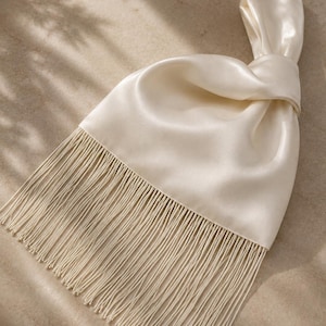 May include: An off-white satin handbag featuring a knotted handle design and a long, straight fringe trim along the bottom edge. The smooth, lustrous fabric creates soft folds around the knot, while the thin, dangling fringe adds texture.