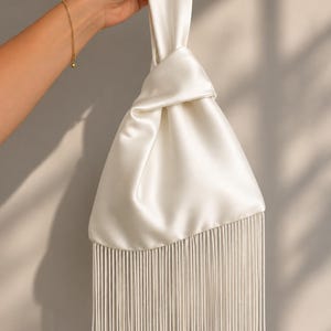 Puede incluir: Un bolso de mano de satén blanco con un diseño de asa superior anudada y un borde de flecos largos y rectos en la parte inferior. El bolso tiene un acabado liso y brillante con forma triangular, y los flecos cuelgan por debajo del cuerpo.