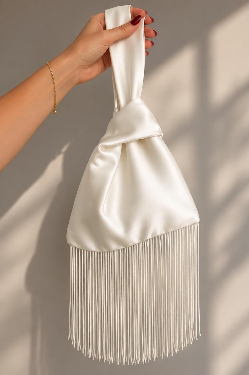 May include: A white satin handbag featuring a knotted top handle design and a long, straight fringe trim along the bottom edge. The bag has a smooth, lustrous finish and a triangular shape, with the fringe extending significantly below the main body.