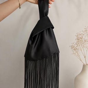 May include: A black satin handbag featuring a knotted handle design and a long, straight fringe trim along the bottom edge. The fabric has a smooth, reflective finish, and the bag is constructed with a single continuous piece of fabric.