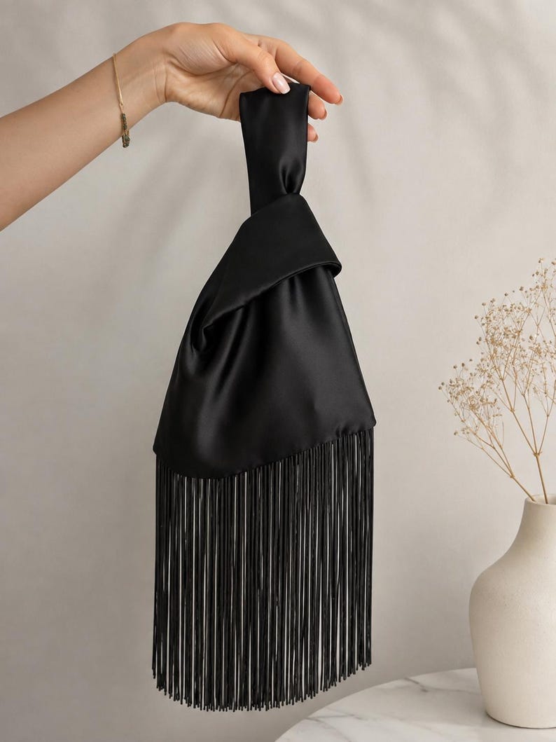May include: A black satin handbag featuring a knotted handle design and a long, straight fringe trim along the bottom edge. The fabric has a smooth, reflective finish, and the bag is constructed with a single continuous piece of fabric.