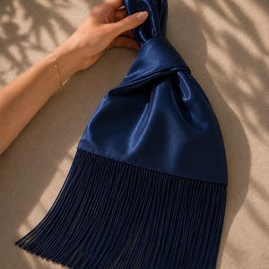 May include: A dark navy blue satin handbag featuring a knotted top handle design and a long, straight fringe trim that extends from the base of the bag. The fabric has a smooth, reflective sheen, and the fringe consists of many thin, vertical strands.