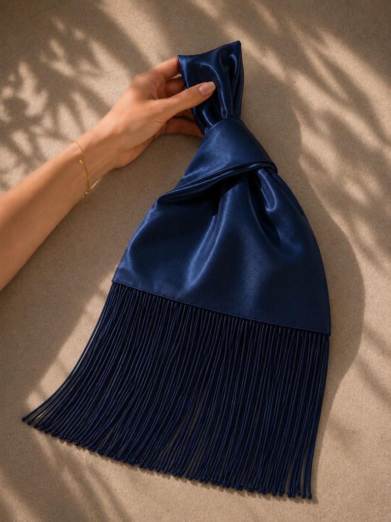 May include: A dark navy blue satin handbag featuring a knotted top handle design and a long, straight fringe trim that extends from the base of the bag. The fabric has a smooth, reflective sheen, and the fringe consists of many thin, vertical strands.