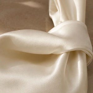 May include: A cream-colored handbag crafted from a smooth, lustrous satin fabric. The bag features a distinctive knotted handle design, creating a soft, draped silhouette with gentle folds and a reflective surface that catches the light.