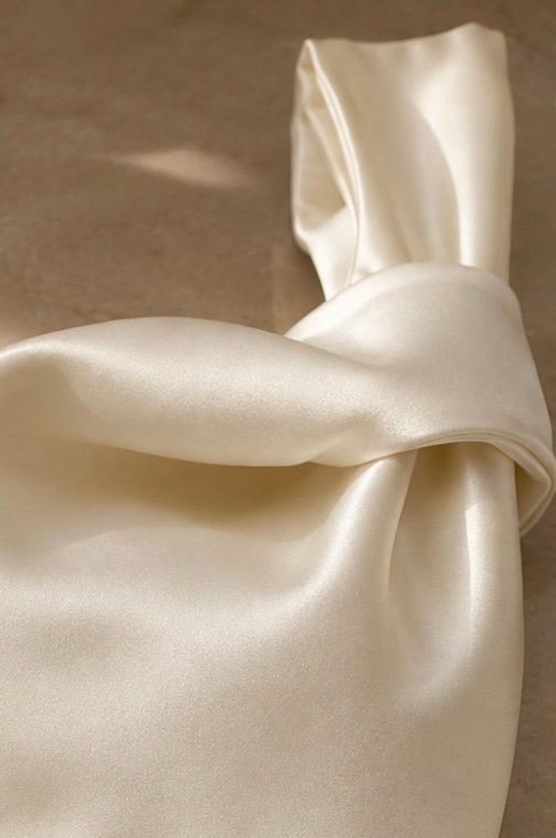 May include: A cream-colored handbag crafted from a smooth, lustrous satin fabric. The bag features a distinctive knotted handle design, creating a soft, draped silhouette with gentle folds and a reflective surface that catches the light.
